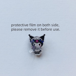 Puede incluir: Un pin de acr&iacute;lico con un personaje de dibujos animados en blanco y negro, un emblema de calavera rosa y un lazo azul. El texto dice: protective film on both side, please remove it before use. Una pel&iacute;cula transparente parcialmente despegada cubre el personaje.