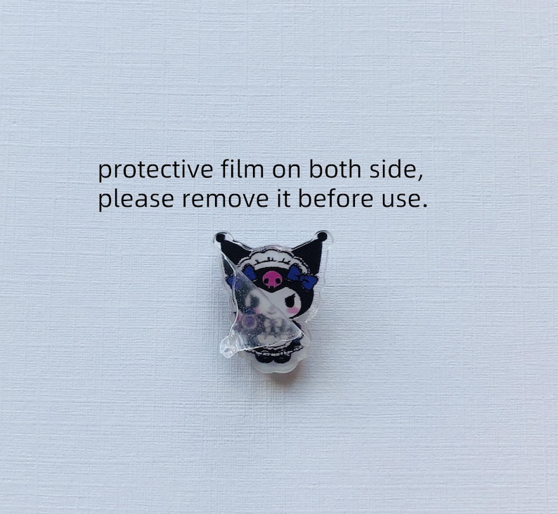 Puede incluir: Un pin de acr&iacute;lico con un personaje de dibujos animados en blanco y negro, un emblema de calavera rosa y un lazo azul. El texto dice: protective film on both side, please remove it before use. Una pel&iacute;cula transparente parcialmente despegada cubre el personaje.