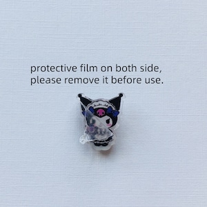 Op de afbeelding: Een heldere acryl bedel met een stripfiguur met zwarte oren, een roze gezicht en blauwe strikken. De bedel heeft een beschermfolie, met de tekst "protective film on both side, please remove it before use."