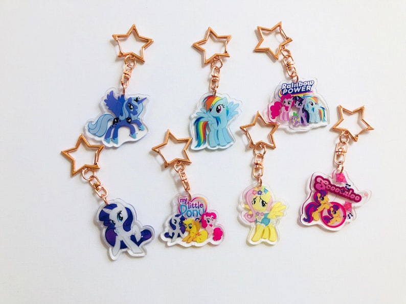 May include: Six My Little Pony keychains with gold star-shaped charms. The keychains feature different characters, including Twilight Sparkle, Rainbow Dash, Pinkie Pie, Fluttershy, Rarity, and Applejack. Each keychain has a clear acrylic charm with a colorful image of the character.