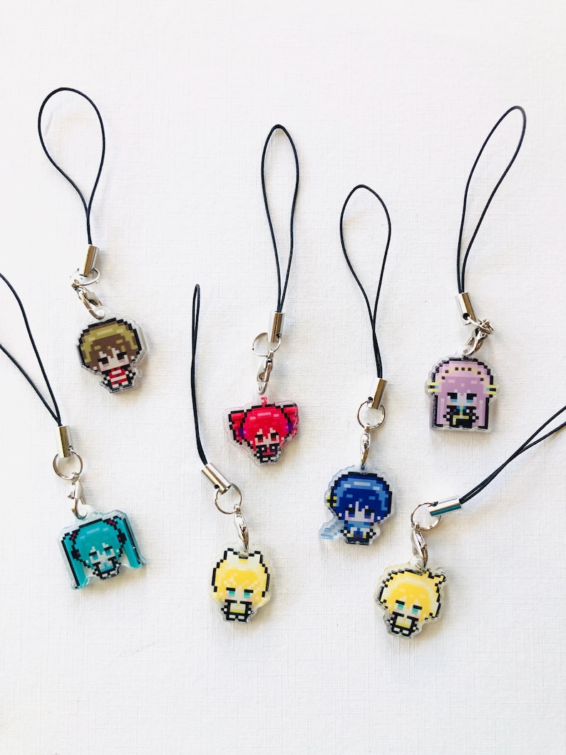 Puede incluir: Una colecci&oacute;n de siete dijes de personajes en pixel art unidos a bucles de cord&oacute;n negro con cierres de metal plateado. Cada dije presenta una figura de estilo anime con colores de cabello distintos, incluyendo turquesa, rubio, rosa, azul y marr&oacute;n, en un dise&ntilde;o de cuadr&iacute;cula.