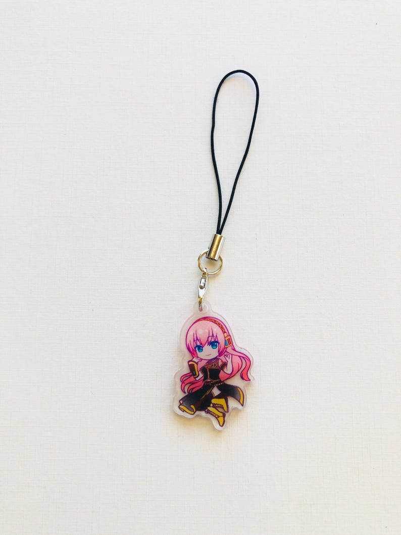 Peut inclure: Un charm en acrylique transparent repr&eacute;sentant un personnage d'anime de style chibi aux cheveux roses et aux yeux bleus, v&ecirc;tu d'une tenue noire et dor&eacute;e. Le charm est attach&eacute; &agrave; un cordon noir avec un fermoir en m&eacute;tal argent&eacute;.