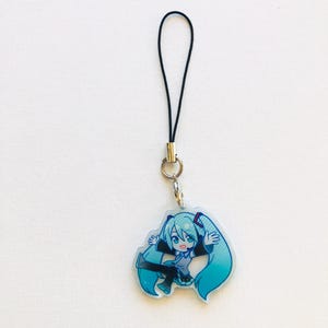 Peut inclure: Un porte-cl&eacute;s avec un personnage de dessin anim&eacute; aux longs cheveux turquoise et une tenue assortie. Le porte-cl&eacute;s est attach&eacute; &agrave; un cordon noir avec un anneau et un fermoir en m&eacute;tal argent&eacute;. Le personnage est dans une pose dynamique.