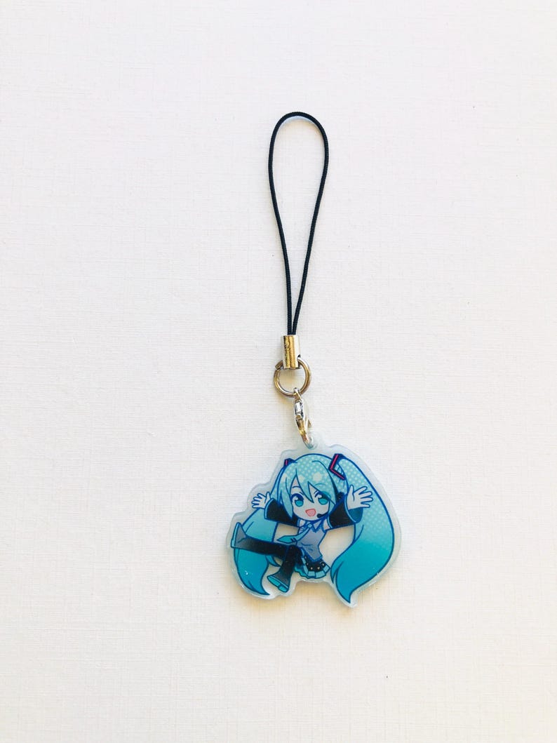 Peut inclure: Un porte-cl&eacute;s avec un personnage de dessin anim&eacute; aux longs cheveux turquoise et une tenue assortie. Le porte-cl&eacute;s est attach&eacute; &agrave; un cordon noir avec un anneau et un fermoir en m&eacute;tal argent&eacute;. Le personnage est dans une pose dynamique.