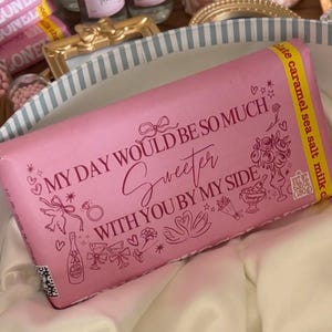 May include: A pink chocolate bar with the text "MY DAY WOULD BE SO MUCH Sweeter WITH YOU BY MY SIDE" and decorative illustrations. The bar is in a striped box with other gifts, including small bottles and pink floral cups.