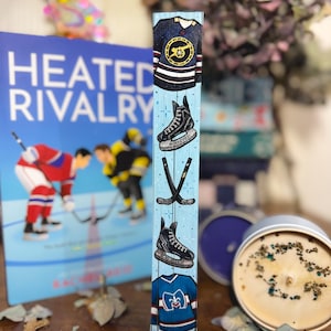 May include: A wooden hockey-themed decorative block with painted images of jerseys, skates, and hockey sticks on a light blue background. A book titled "Heated Rivalry" is visible in the background. A candle in a tin is also in the image.