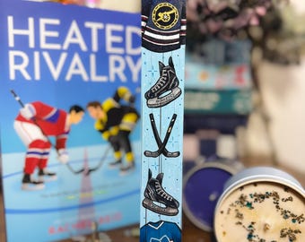 May include: A wooden hockey-themed decorative block with painted images of jerseys, skates, and hockey sticks on a light blue background. A book titled "Heated Rivalry" is visible in the background. A candle in a tin is also in the image.
