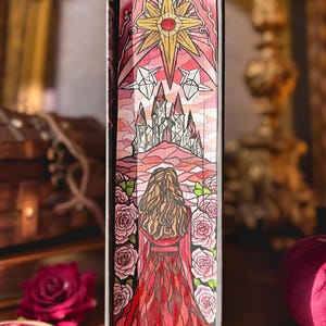 Könnte beinhalten: Buchrücken mit farbenfroher Illustration. Das Kunstwerk zeigt eine Frau in einem roten Kleid, ein Schloss und einen großen Stern. Rosa Rosen umrahmen die Frau. Die Farbpalette umfasst Rot, Rosa und Gold.