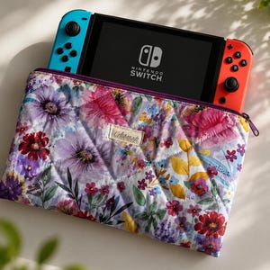 May include: A floral quilted case with a Nintendo Switch console inside. The case features a vibrant pattern of purple, pink, and yellow flowers on a white background. The Nintendo Switch has blue and red controllers. The case has a purple zipper and a small label.