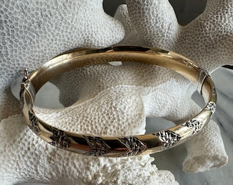 Könnte beinhalten: Ein goldenes Armreif-Armband mit geometrischem Muster. Das Armband ist aus zweifarbigem Metall gefertigt, mit einer goldenen Außenseite und einer silbernen Innenseite.