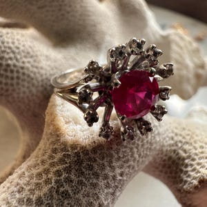 Op de afbeelding: Een zilveren ring met een grote, ronde, robijnrode edelsteen in het midden. De ring heeft een uniek ontwerp met kleine, sprankelende accenten rondom de centrale steen, die op een sterrenuitbarsting lijken. De ring wordt gepresenteerd op een getextureerd, gebroken witte ondergrond.