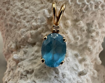 Puede incluir: Un colgante de oro con una piedra preciosa ovalada de color azul claro. La piedra preciosa está sujeta por cuatro pequeñas garras. El colgante tiene una fianza de oro para una cadena. La piedra preciosa tiene una superficie facetada.