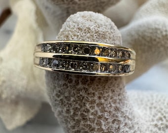 May include: A gold-toned ring with two rows of small, dark gemstones. The ring has a classic band design and is set against a textured, off-white background, highlighting the ring's sparkle and detail.