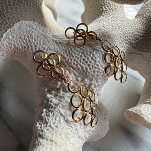 May include: A pair of gold earrings with a delicate, circular design. The earrings are made of multiple interconnected circles, creating a cascading effect. The circles are hollow and have a slightly textured surface.