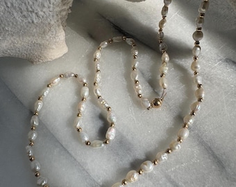 May include: A delicate pearl necklace with gold-colored beads and a clasp. The necklace features irregularly shaped white pearls, strung together with small gold beads. The clasp is also gold-colored. The necklace is displayed on a white marble surface.