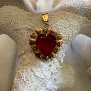 Könnte beinhalten: Goldfarbener Anhänger mit einem großen, herzförmigen, roten Edelstein. Der Edelstein ist von kleinen goldenen Herzen umgeben. Der Anhänger hängt an einer kleinen, goldenen Öse.