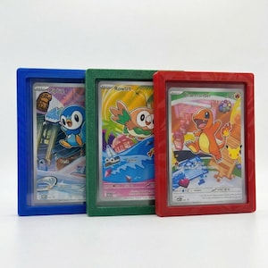 May include: Three framed Pokémon trading cards. The frames are blue, green, and red. The cards feature the characters Piplup, Rowlet, and Charmander. The Piplup card has a cityscape background. The Rowlet card has a tropical background. The Charmander card has a pixelated background.