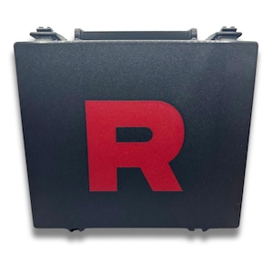 May include: Black rectangular case with a red letter "R" on the front. The case has a handle and latches on top. The case is likely used for storage or transport.