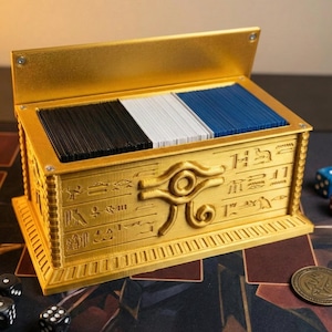 May include: A gold-colored card storage box with an open lid, filled with black, white, and blue cards. The box features hieroglyphic-style designs and an eye symbol on the front. Dice and a coin are visible.