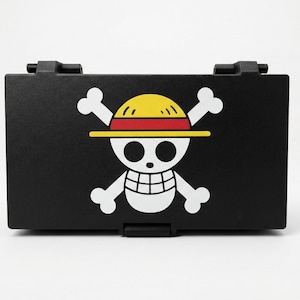 Może przedstawiać: Czarny, prostokątny pojemnik z symbolem Jolly Roger z anime One Piece. Symbol zawiera białą czaszkę z incydentalnymi kośćmi, ż&oacute;łty słomkowy kapelusz oraz czerwone i ż&oacute;łte akcenty. Pojemnik ma klapę na zawiasach.