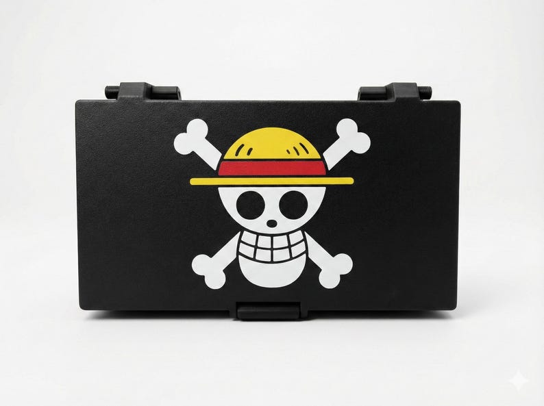 Może przedstawiać: Czarny, prostokątny pojemnik z symbolem Jolly Roger z anime One Piece. Symbol zawiera białą czaszkę z incydentalnymi kośćmi, ż&oacute;łty słomkowy kapelusz oraz czerwone i ż&oacute;łte akcenty. Pojemnik ma klapę na zawiasach.