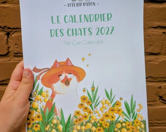 Può includere: Un calendario di carta bianca intitolato LE CALENDRIER DES CHATS 2027 e The Cat Calendar, con l'illustrazione di un gatto rosso che indossa un cappello di paglia in un campo di fiori gialli e foglie verdi con piccole api e stelle gialle.