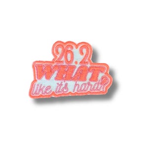 Puede incluir: Un parche bordado de color coral con el texto "26.2 WHAT like its hard?" en una fuente lúdica y estilizada. El parche tiene un fondo blanco y un efecto de sombra, lo que sugiere un diseño en relieve. El parche es probablemente para corredores de maratón.