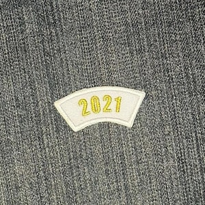 May include: A white patch with a curved top sewn onto a denim fabric background. The patch has the year 2021 embroidered in gold thread.