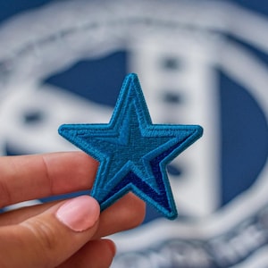 Puede incluir: Un parche azul en forma de estrella con un contorno blanco. El parche se sostiene en una mano contra un fondo borroso.