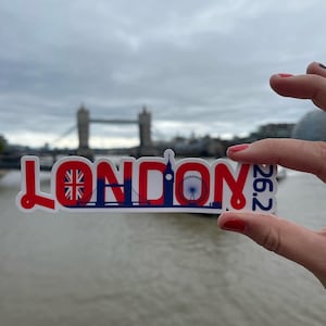 Puede incluir: Una pegatina blanca con la palabra "LONDON" en letras mayúsculas rojas, con una bandera de la Union Jack y monumentos de Londres. El número "26.2" está impreso en azul. La pegatina se sostiene frente a un fondo borroso del Tower Bridge.