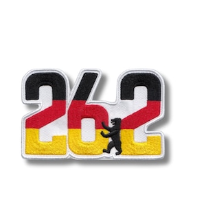 Puede incluir: Parche bordado con el número "262" en los colores de la bandera alemana: negro, rojo y amarillo, con fondo blanco. Una silueta de oso negro está en el centro. Para ropa o accesorios.