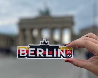 May include: A black and white sticker with the word "BERLIN" in bold white letters on a black background. The sticker has a yellow and red stripe at the top and bottom, representing the German flag. The sticker is held in a hand in front of a blurred image of the Brandenburg Gate.
