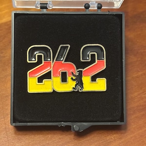 May include: A pin in a clear case, featuring the number "26.2" in black, red, and yellow, representing the German flag. A black bear silhouette is between the numbers. The pin is set against a black background.