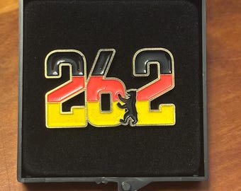 May include: A pin in a clear case, featuring the number "26.2" in black, red, and yellow, representing the German flag. A black bear silhouette is between the numbers. The pin is set against a black background.