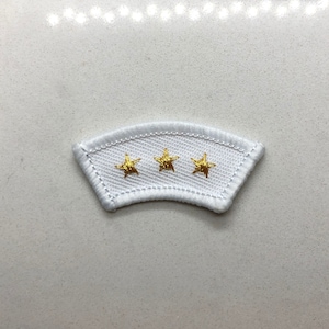 May include: White fabric patch with three gold embroidered stars. The patch is curved and has a white stitched border.