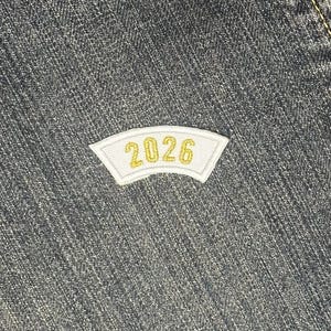 May include: A white patch with gold trim and the number "2026" in gold lettering. The patch is set against a textured, dark denim background.