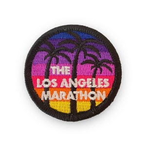 May include: A round, black embroidered patch with a sunset scene and palm trees. The text "THE LOS ANGELES MARATHON" is embroidered in white.