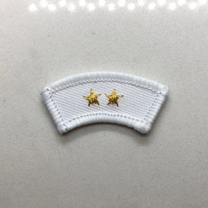 May include: White fabric patch with two gold embroidered stars. The patch is shaped like a curved rectangle.