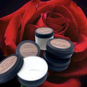 May include: Several small, black tins with wooden-effect lids, some open to show a white, creamy substance. The text "Rivers Edge" and "Grass Fed Tallow" is visible on the lids. A large, red rose provides a soft, blurred background.