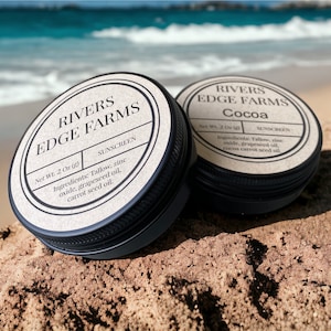 Puede incluir: Dos botes pequeños, redondos, de color marrón con tapas negras. Los botes están etiquetados con "Rivers Edge Farms" y contienen protector solar. El bote de la izquierda está etiquetado con "Sunscreen" y el bote de la derecha está etiquetado con "Cocoa". Ambos botes están sobre una playa de arena.