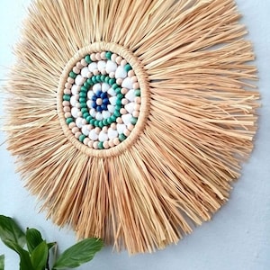 May include: A round, natural fiber wall hanging with a sunburst design. The center features a circular pattern of white, green, and blue beads, surrounded by a woven border. The outer edge has radiating strands of straw.