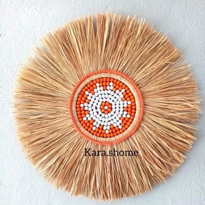 May include: A round, natural fiber wall hanging with a sunburst design. The center features a beaded pattern in orange and white, framed by a coral-colored rope. The text "Kara.shome" is printed at the bottom.