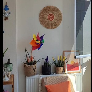 May include: A bright, sunlit interior scene featuring a variety of decorative elements. A colorful pinwheel stands out, alongside potted plants and a framed art print. A woven wall hanging and an orange pillow add warmth.