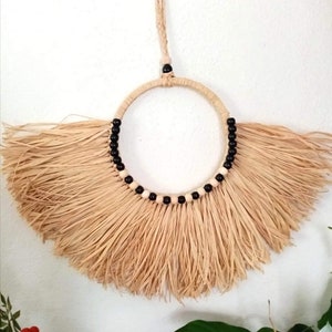 May include: A natural fiber wall hanging with a circular design. The center is a woven ring adorned with black and white beads. The bottom features a fan of long, beige fibers, creating a textured, bohemian aesthetic. A woven loop is at the top for hanging.