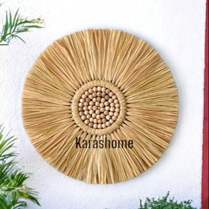 May include: A round, natural-toned wall hanging with a sunflower design. The center features small, light-colored wooden beads, surrounded by radiating, textured straw-like material. The text "Karashome" is printed across the bottom. This decorative piece is suitable for home decor.