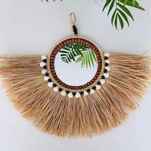 May include: A round wall mirror with a natural, bohemian design. The mirror is framed with a ring of wooden beads and adorned with seashells. The outer edge features a fan of dried straw, creating a textured, decorative piece.