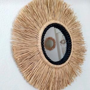 May include: Round wall mirror with a natural raffia fringe. The mirror has a black beaded border and a reflective surface. The raffia is a light tan color, creating a bohemian or coastal aesthetic. The mirror is mounted on a white wall.