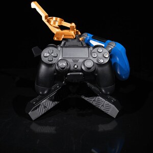 May include: A black PlayStation 4 controller with a gold metal stand and a blue controller on the side. The stand has a textured surface.