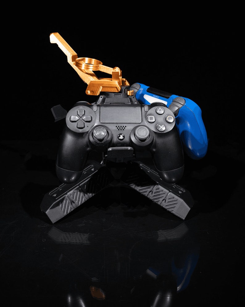May include: A black PlayStation 4 controller with a gold metal stand and a blue controller on the side. The stand has a textured surface.