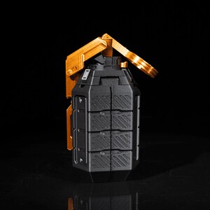 May include: A black and gold grenade with a textured surface and a gold lever. The grenade is on a black surface.